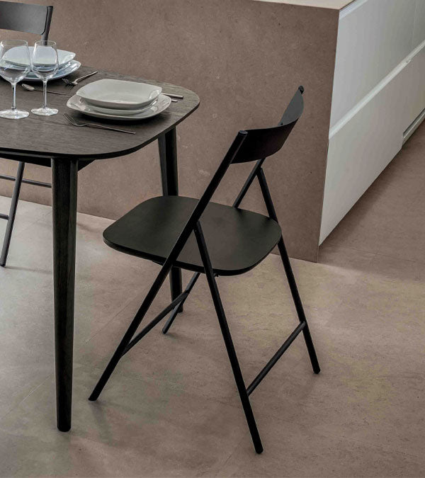  Joe black folding dining chair next to a round dining table with plates and glasses on a neutral background