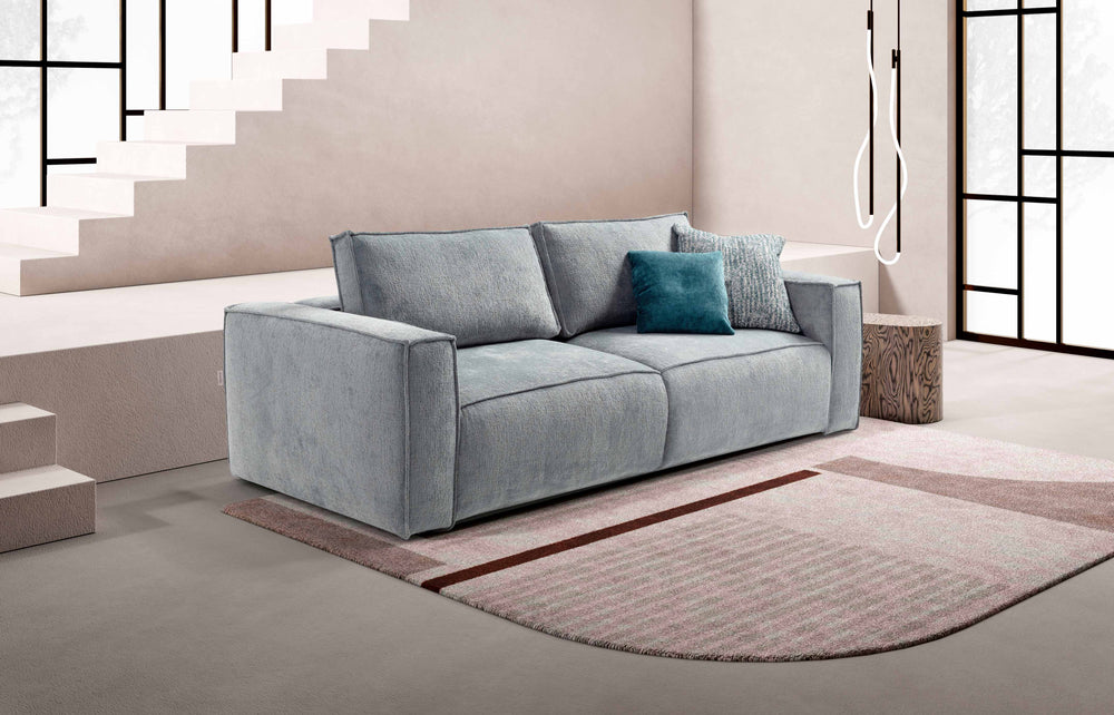 Lush Grey sofa with a blue pillow on a rug in a modern room with stairs and large windows.
